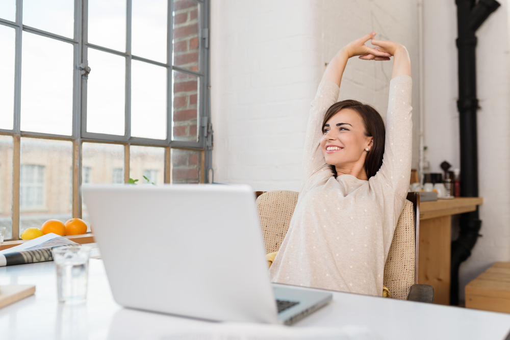 Happy relaxed young woman engaging in remote work in her kitchen with a laptop in front of her stretching her arms above her head and looking out of the window with a smile